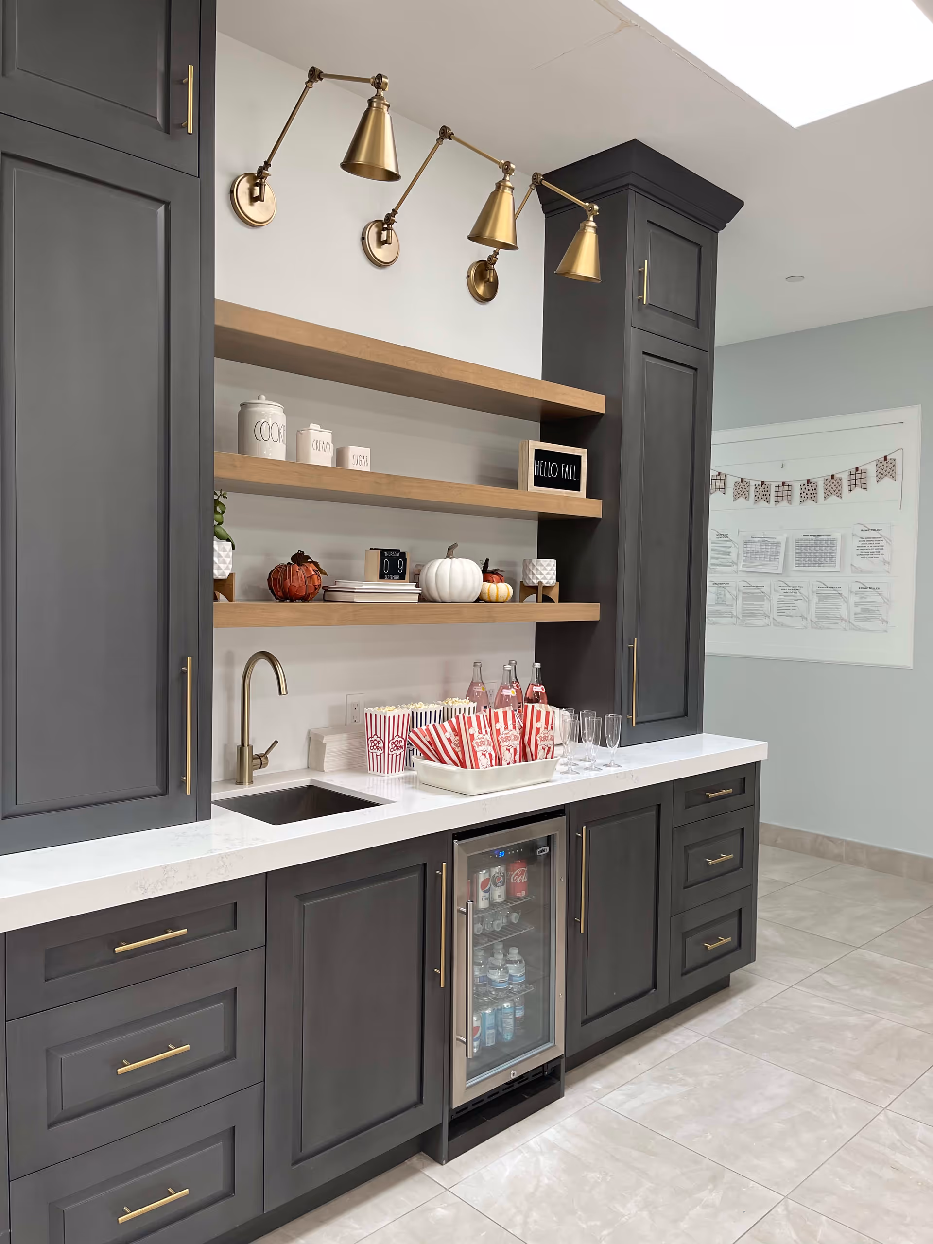 A modern kitchenette area with dark gray cabinets and brass handles, white countertop, and a small sink with a brass faucet. Above the counter are three wooden floating shelves decorated with white jars labeled 'COOK', 'CINNAMON', and 'SUGAR', small pumpkins, books, and a sign that says 'HELLO FALL'. Below the counter is a small beverage refrigerator stocked with drinks. On the counter are popcorn containers, glass bottles with pink liquid, and empty glasses. The floor is tiled in light beige, and a bulletin board with papers and a decorative banner is visible on the light blue wall in the background.