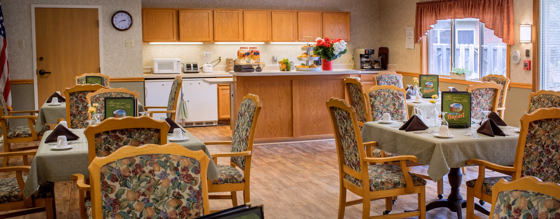 A dining room in a senior living facility with several tables covered with green tablecloths, each set with cups, napkins, and menus. The chairs have wooden frames with floral upholstered cushions. In the background, there is a kitchenette area with wooden cabinets, a microwave, a coffee maker, and a small refrigerator. A window with a red valance lets in natural light.