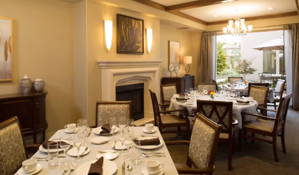 Elegant dining room with round tables set with white tablecloths, plates, glasses, and brown napkins. The room features upholstered chairs, a fireplace with wall sconces and artwork above it, a sideboard with decorative vases, and a large window with curtains looking out to a patio with outdoor seating and greenery.