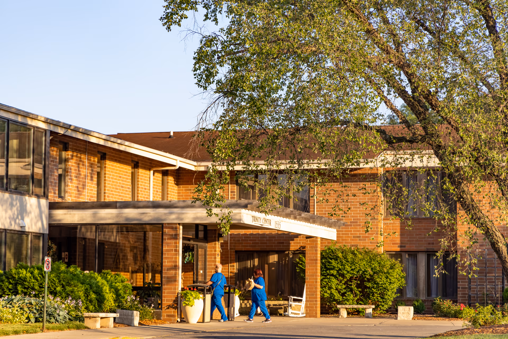 Exterior view of a brick senior living facility building with large windows and a covered entrance. Two people in blue scrubs are walking near the entrance. There are trees and bushes around the building, and the scene is lit by warm sunlight.