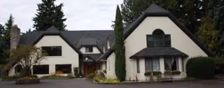Large Tudor-style residential building with steep gabled roofs, white exterior walls, and a circular driveway surrounded by trees.