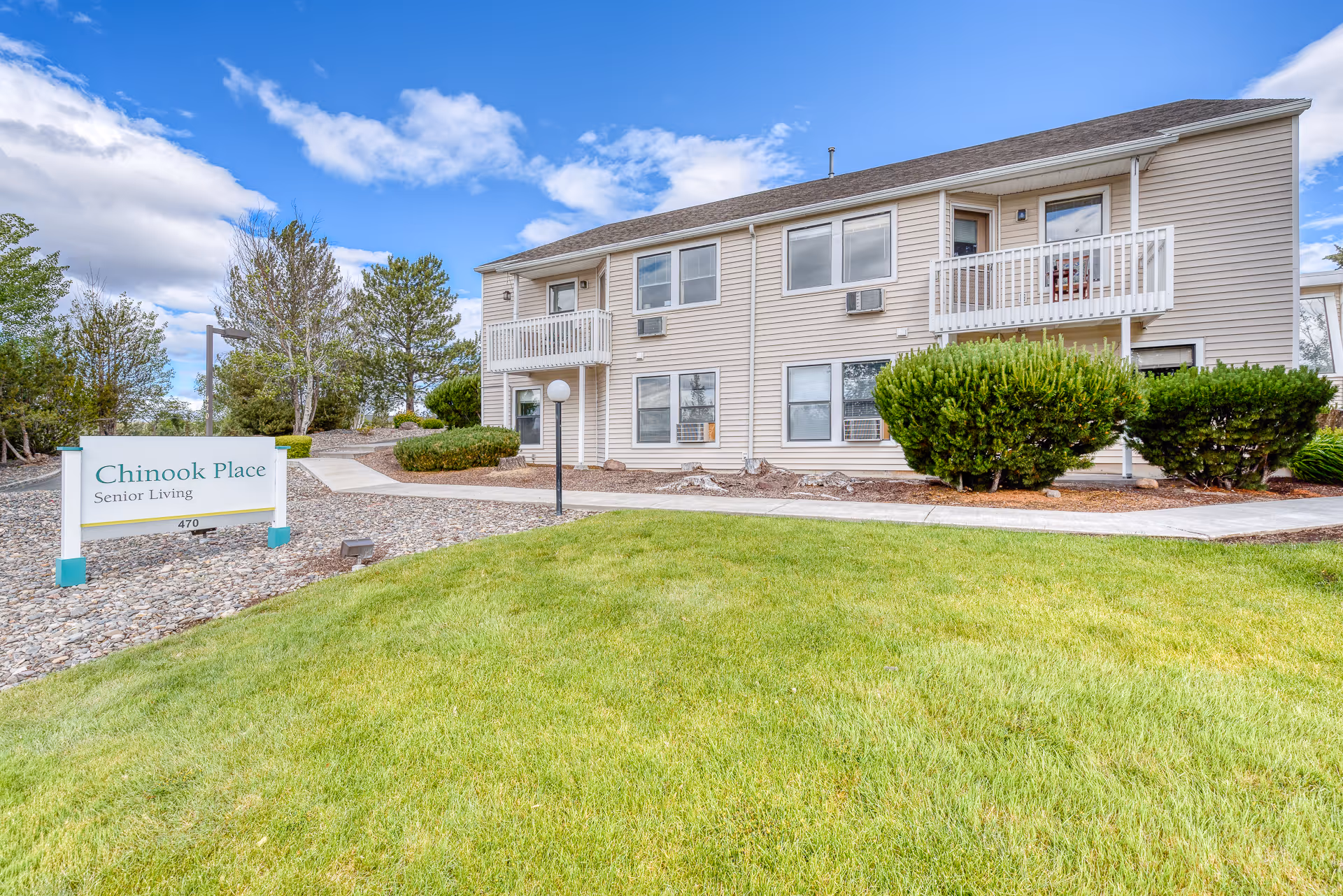 Front exterior of a two-story senior living building with balconies, a lawn, and a sign reading "Chinook Place Senior Living".