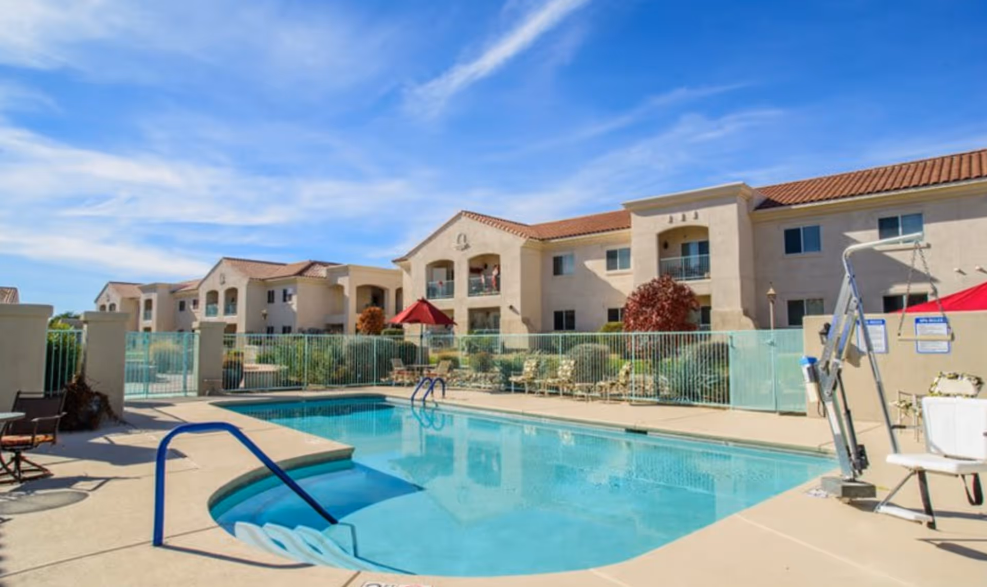 Outdoor swimming pool with lounge chairs and a fenced deck in front of a two-story apartment-style senior living building under a blue sky.