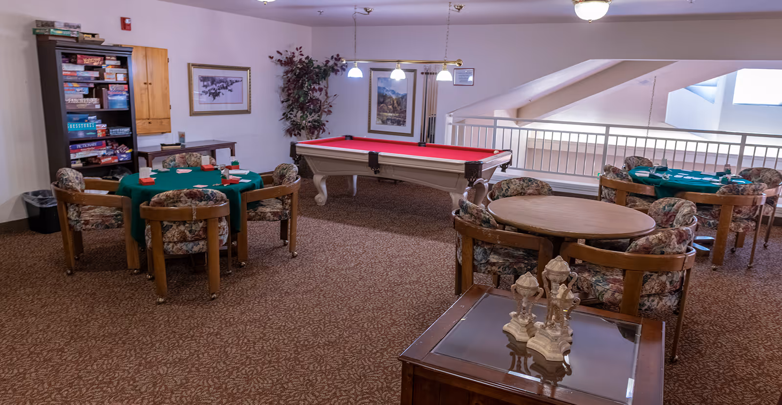 A recreational room in a senior living community featuring a red pool table, several round tables with floral upholstered chairs, and a bookshelf filled with board games. The room has carpeted flooring, framed pictures on the walls, and a railing overlooking a lower level.