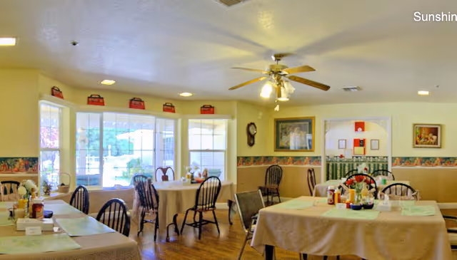 Bright communal dining room with several tables and chairs, a bay window, ceiling fan, and wall decorations.