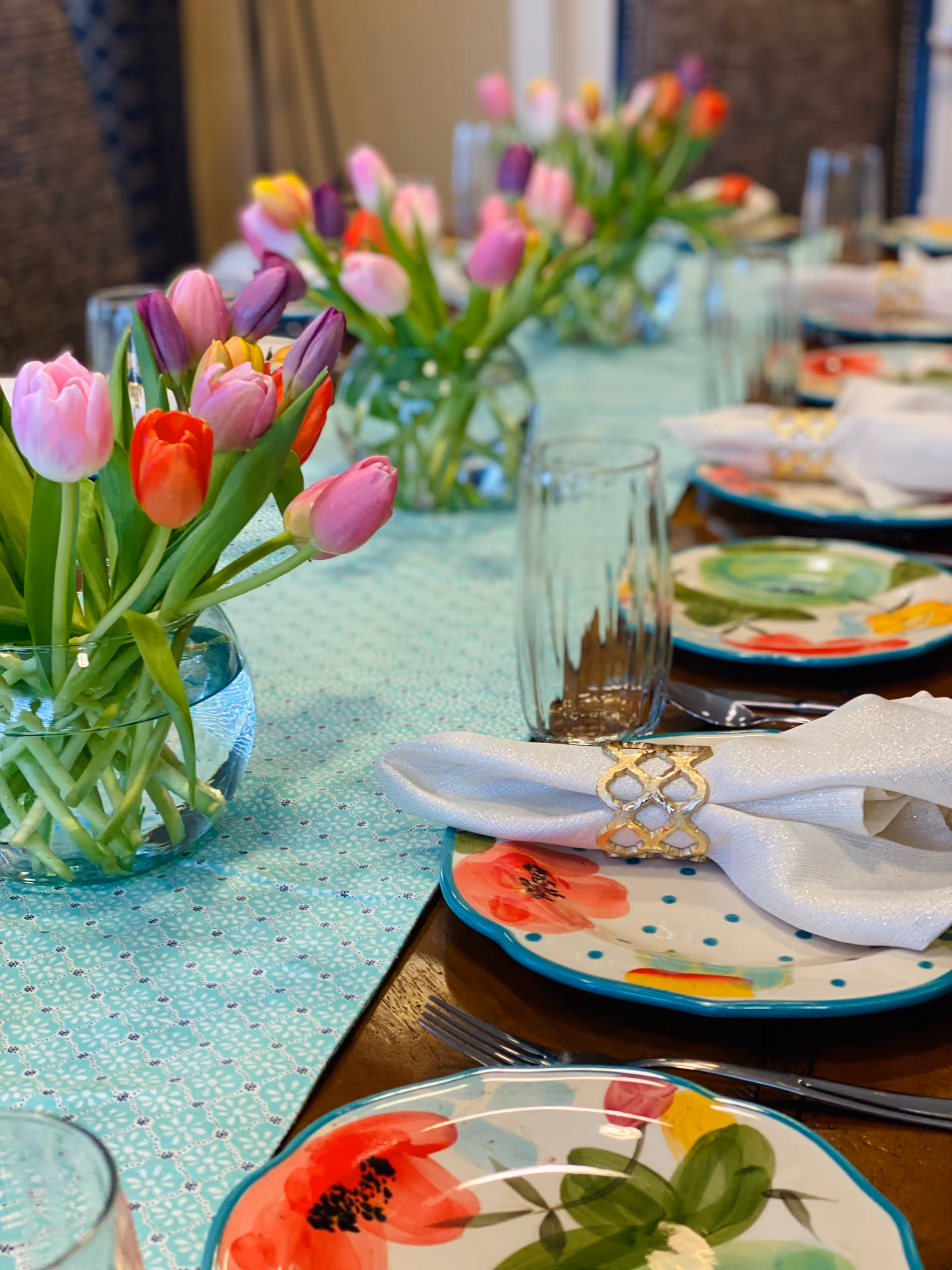 A dining table set with colorful floral-patterned plates, white napkins with gold napkin rings, clear drinking glasses, and vases filled with vibrant tulips in various colors including pink, red, purple, and yellow. The table has a light blue patterned table runner.