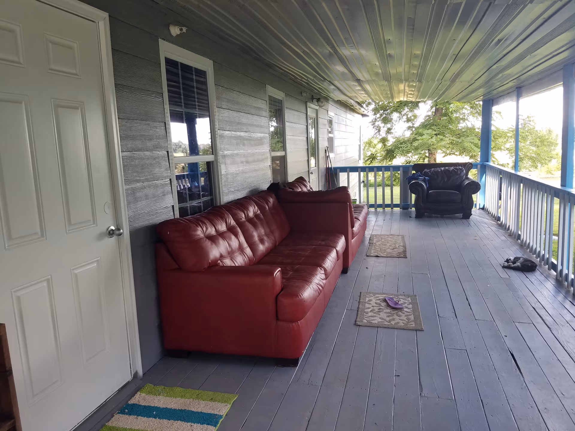 Covered outdoor porch area with a red leather sofa, a matching red leather loveseat, and a black armchair. The porch has a gray wooden floor with two small rugs and a single purple slipper. There is a white door on the left side and windows along the wall. Trees and greenery are visible beyond the porch railing.