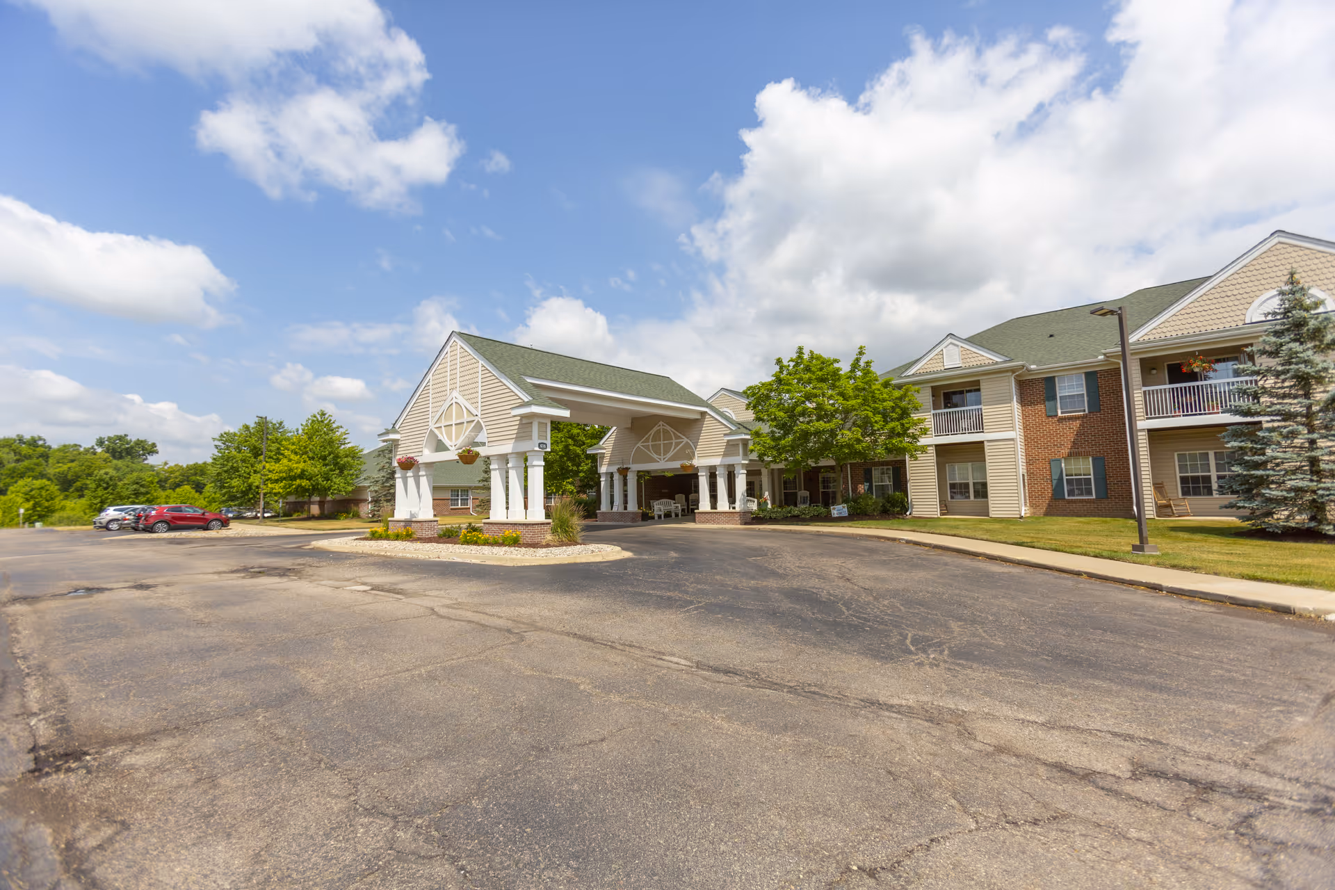 Exterior view of Independence Village of Grand Ledge senior living facility showing a large covered entrance with columns, a parking lot with several cars, and a two-story building with balconies and greenery under a partly cloudy sky.