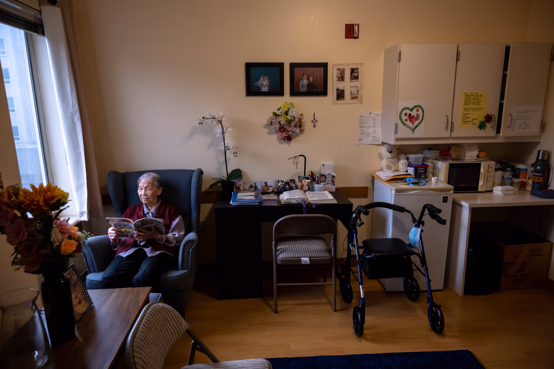 An elderly woman sits in a blue armchair near a window, reading a magazine in a cozy room. The room has a wooden floor, a table with flowers, a desk with photos and papers, a walker, and a kitchenette area with cabinets, a microwave, and a small refrigerator. The walls are decorated with framed photos and a floral wreath.