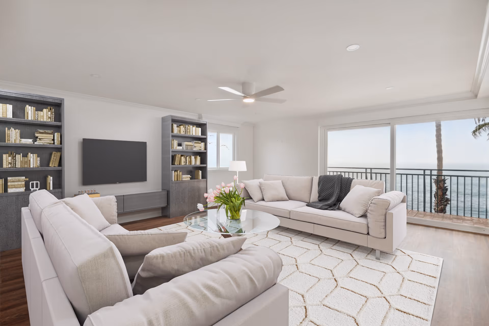 A bright and modern living room with two light-colored sofas arranged around a glass coffee table with a vase of pink flowers. There are two tall bookshelves filled with books flanking a wall-mounted flat-screen TV. Large sliding glass doors open to a balcony with a view of the ocean and palm trees. The room has a white ceiling fan and a light-colored geometric patterned rug on wooden flooring.