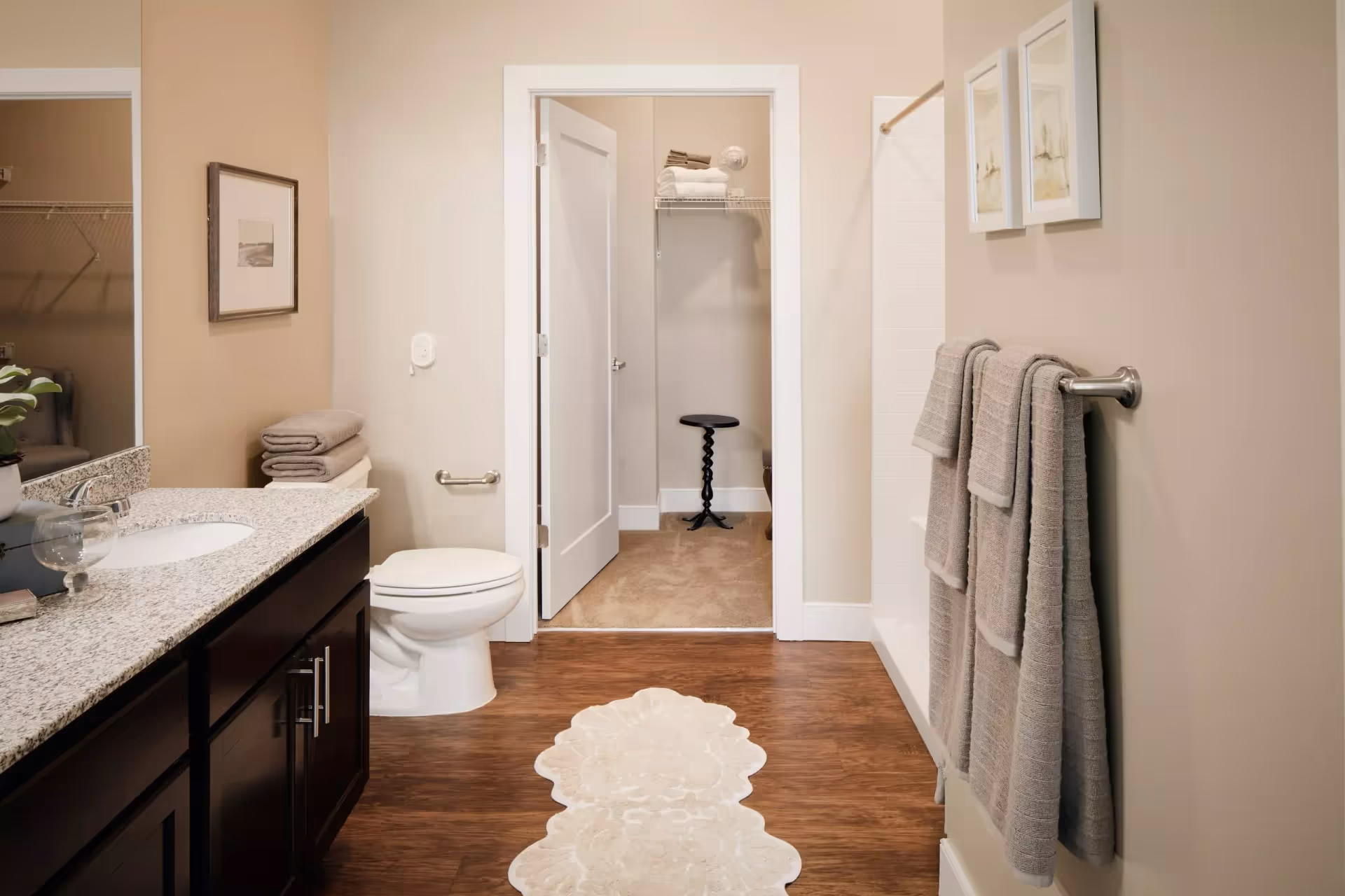 A bathroom with a granite countertop vanity on the left, a white toilet next to it, and a towel rack with three gray towels on the right wall. There is a white door in the center leading to a small closet with shelves holding folded towels and a small black stool. The floor is wood with a decorative beige rug in the middle.