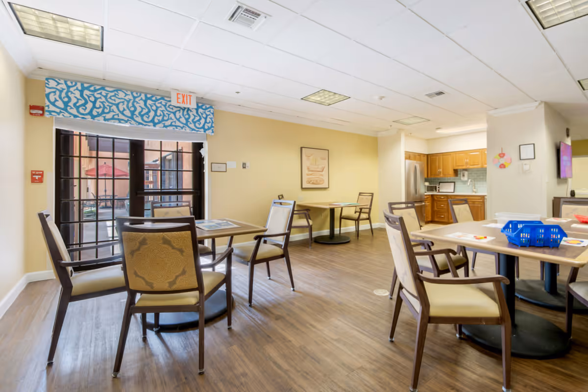 A bright and clean common area in a senior living facility with several tables and chairs arranged for seating. The room has wood flooring, beige walls, and a large window with a blue patterned valance and an exit sign above it. In the background, there is a kitchen area with wooden cabinets, a stainless steel refrigerator, and a microwave. The room is well-lit with ceiling lights.