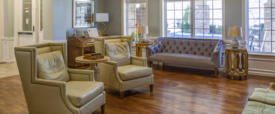 A bright and spacious living room area in an assisted living facility featuring two beige leather armchairs with nailhead trim, a tufted gray sofa, wooden side tables with lamps, a piano against the wall, and large windows letting in natural light.