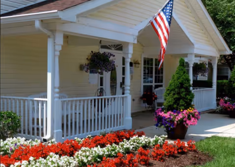 Front porch of a yellow building with white railings and columns, decorated with hanging flower baskets and potted plants. An American flag is displayed on the porch. There are vibrant red and white flowers planted in the garden bed in front of the porch.