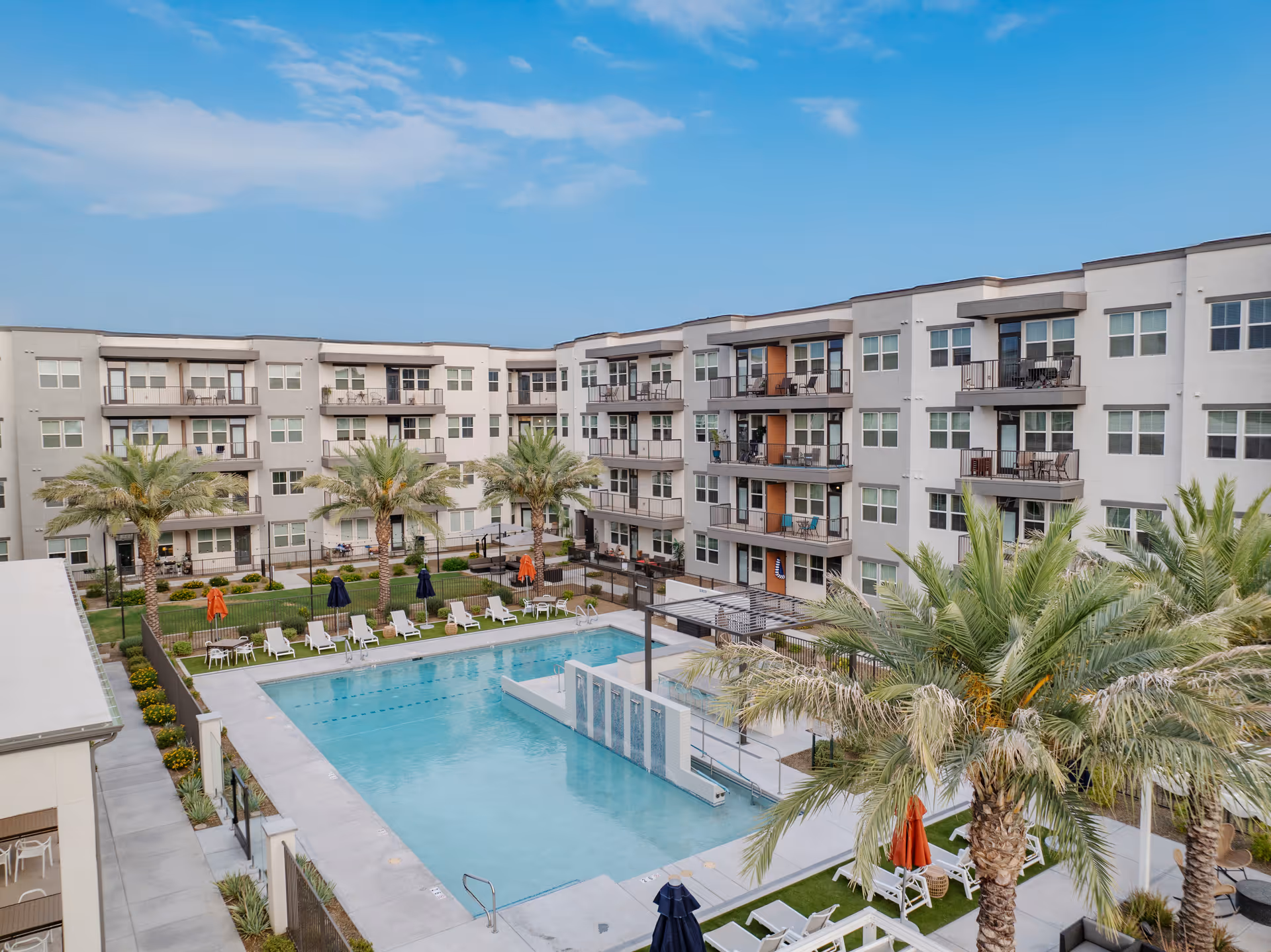 View of a four-story apartment-style courtyard with a swimming pool, palm trees, lounge chairs, and surrounding balconies.