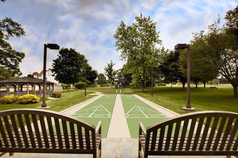 Two shuffleboard courts with wooden benches, lamp posts, a gazebo, and trees in a landscaped outdoor park area.