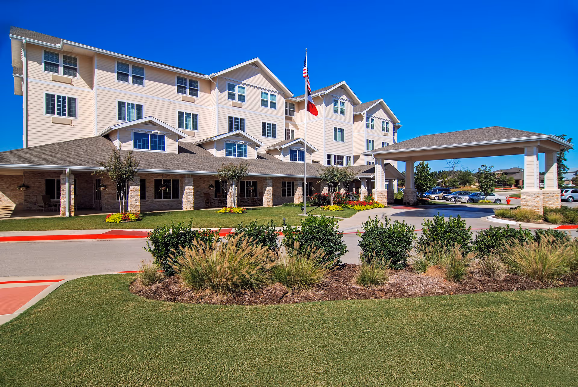 Exterior view of a multi-story senior living facility with beige siding and stone accents. The building has multiple windows and a covered entrance with a driveway. There is a flagpole with two flags in front of the building, surrounded by landscaped bushes and grass under a clear blue sky.