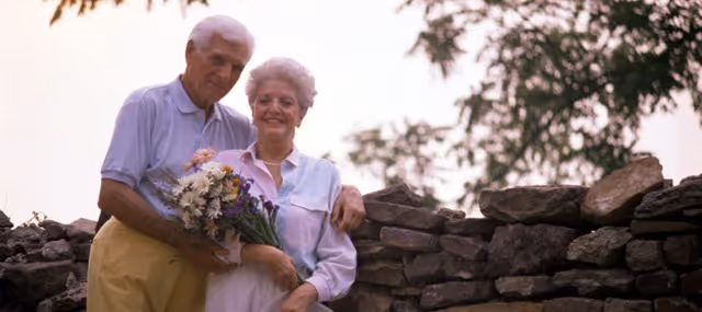 An elderly couple standing outdoors in front of a stone wall. The man is holding a bouquet of flowers and has his arm around the woman, who is smiling. Trees and foliage are visible in the background.