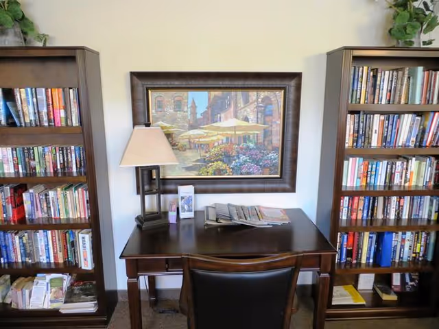 A cozy reading or study area with a dark wooden desk and a black chair. On the desk, there is a table lamp, some brochures, and a display of magazines or pamphlets. Behind the desk, a framed painting of an outdoor café scene with umbrellas and flowers hangs on the wall. On either side of the desk, there are tall wooden bookshelves filled with books and some additional reading materials at the bottom.