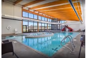 Indoor swimming pool area with large windows letting in natural light, a hot tub adjacent to the pool, a red slide leading into the pool, and seating around the pool deck. The ceiling has wooden beams and the pool area has a clean, modern design.