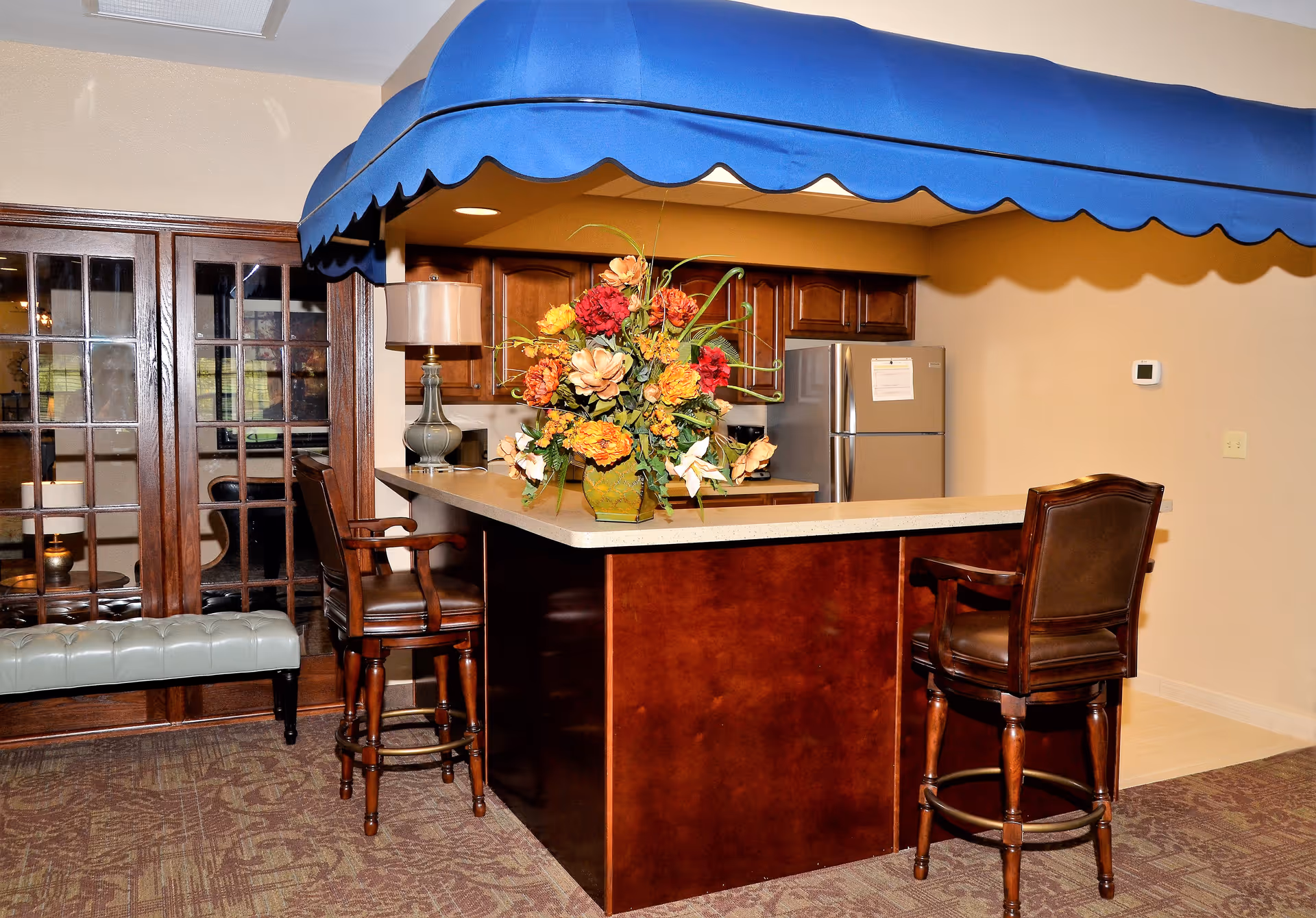 Interior view of a kitchen area with a wooden island counter topped with a beige countertop. Two high wooden chairs with leather seats are positioned at the counter. A large floral arrangement in a green vase sits on the counter. Above the counter is a blue scalloped canopy. In the background, there are wooden cabinets and a stainless steel refrigerator. To the left, there are glass-paneled double doors and a cushioned bench.
