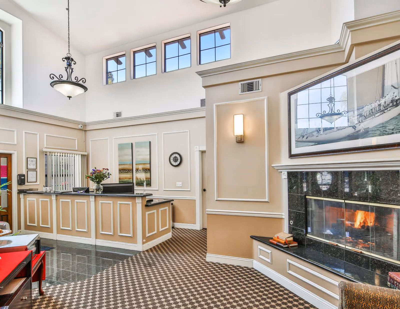 Interior view of a reception area in a senior living facility with a front desk, decorative wall paneling, a lit fireplace with a large framed picture above it, a hanging light fixture, and multiple windows near the ceiling allowing natural light.