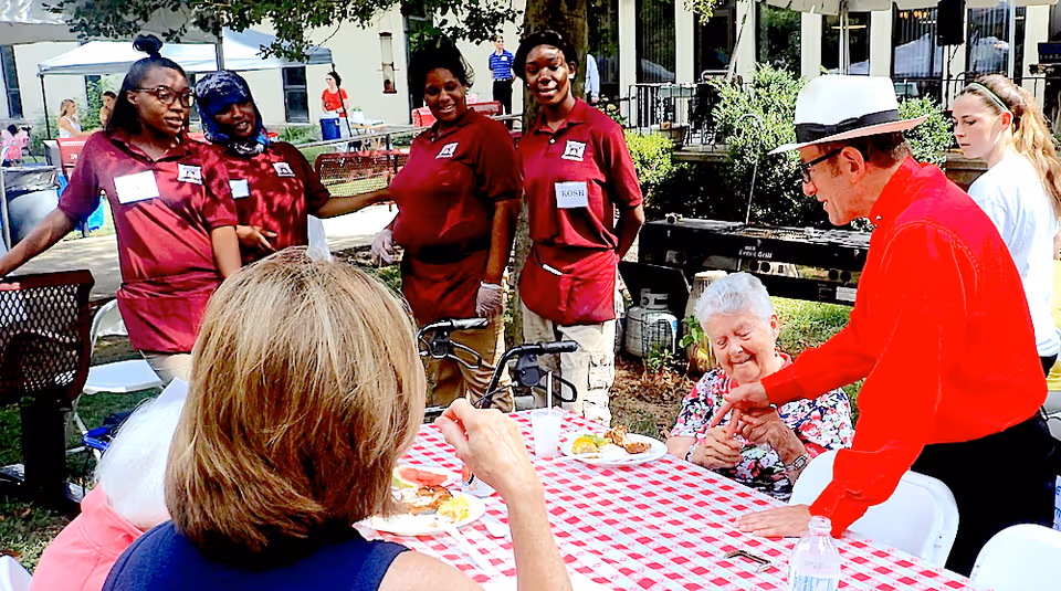 A group of staff and elderly residents gathered around a red-and-white checkered picnic table outdoors during a community event.