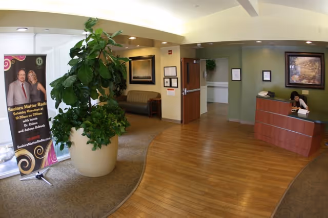 Reception area of Upland Rehabilitation & Care Center featuring a wooden reception desk with a woman seated behind it, a large potted plant, a banner advertising Seniors Matter Radio, a seating area with a couch, framed artwork on the walls, and a combination of carpet and wooden flooring.