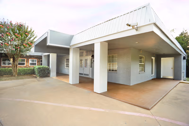 Exterior view of a single-story building with a covered entrance supported by white pillars. The building has gray walls, several windows, and a white door. There is a concrete driveway and a small landscaped area with bushes and a flowering tree on the left side.
