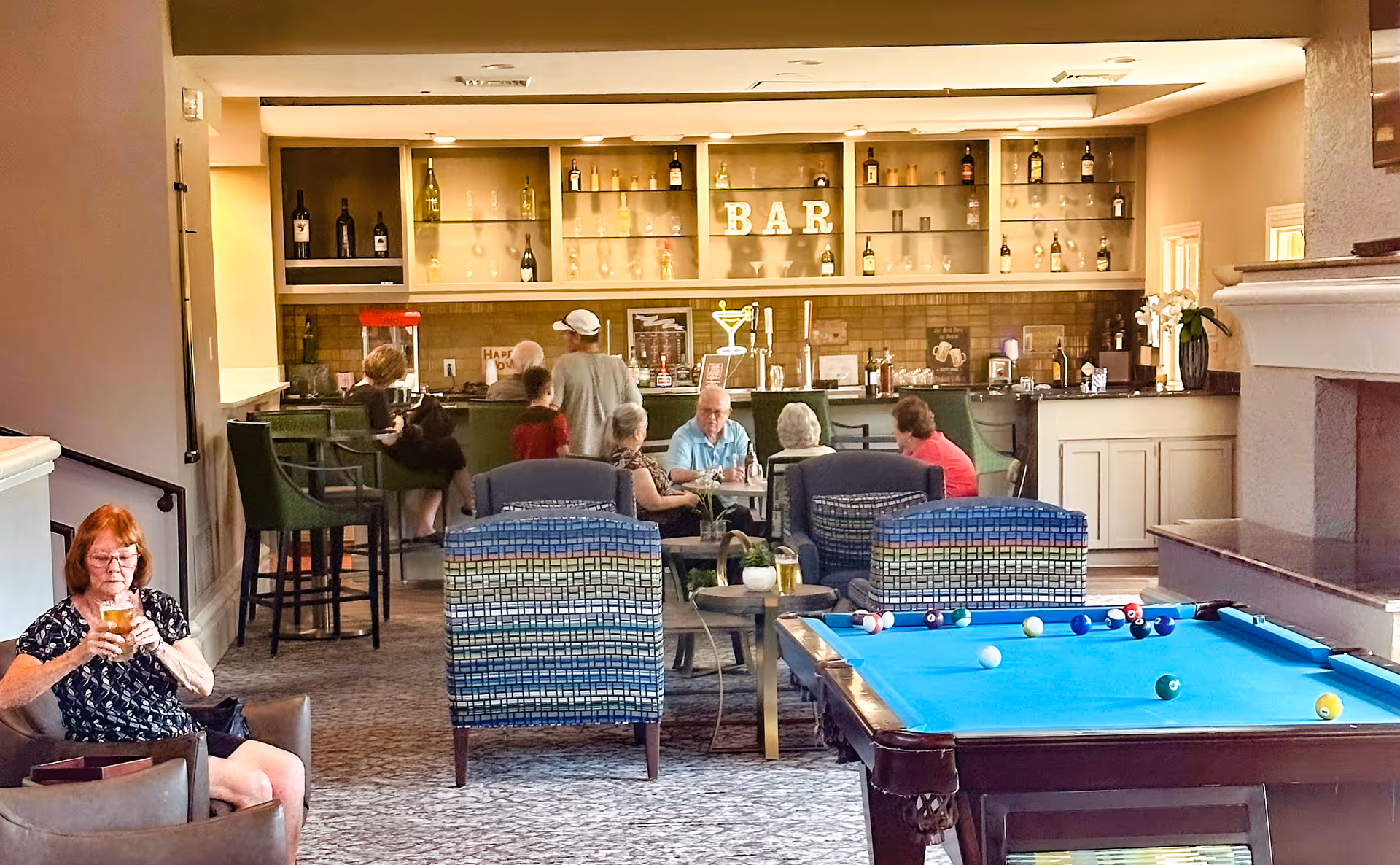 Residents socialize in a senior living lounge with a bar, seating area, and a pool table in the foreground.