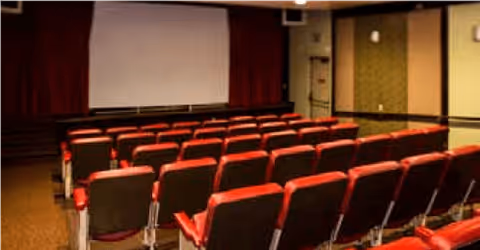 Interior view of a small theater or presentation room with rows of red and black cushioned seats facing a large white projection screen. The room has beige walls and a dark carpeted floor.