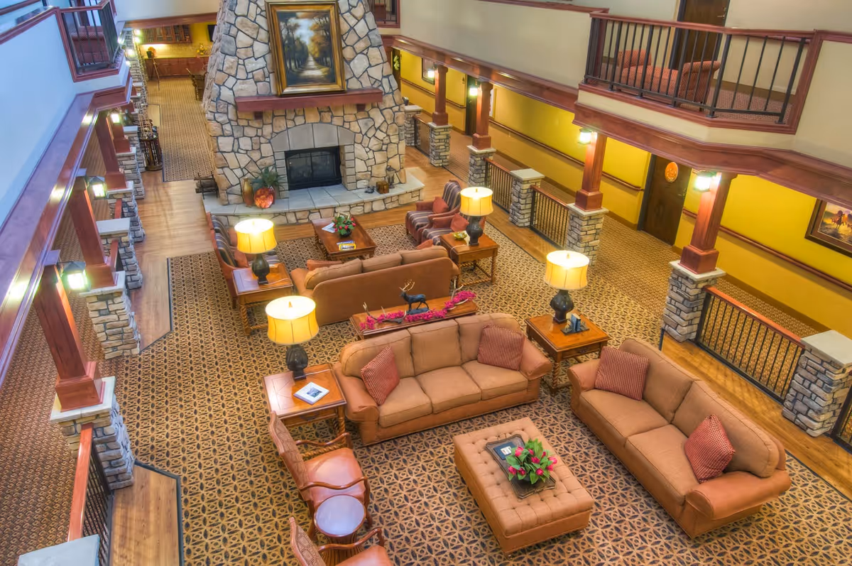 Spacious senior living facility common area with multiple brown sofas and armchairs arranged around a central ottoman and coffee tables. The room features a large stone fireplace with a painting above it, warm lighting from table lamps, and patterned carpet flooring. The area is surrounded by a balcony with wooden railings and stone pillars.
