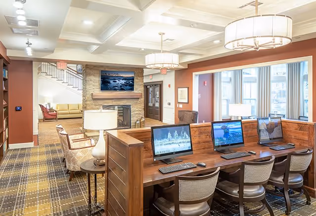 A well-lit senior living facility common area featuring a row of four computer workstations with chairs, a stone fireplace with a mounted TV above it, comfortable seating including armchairs and sofas, and large windows letting in natural light. The room has warm earth tones and modern light fixtures.