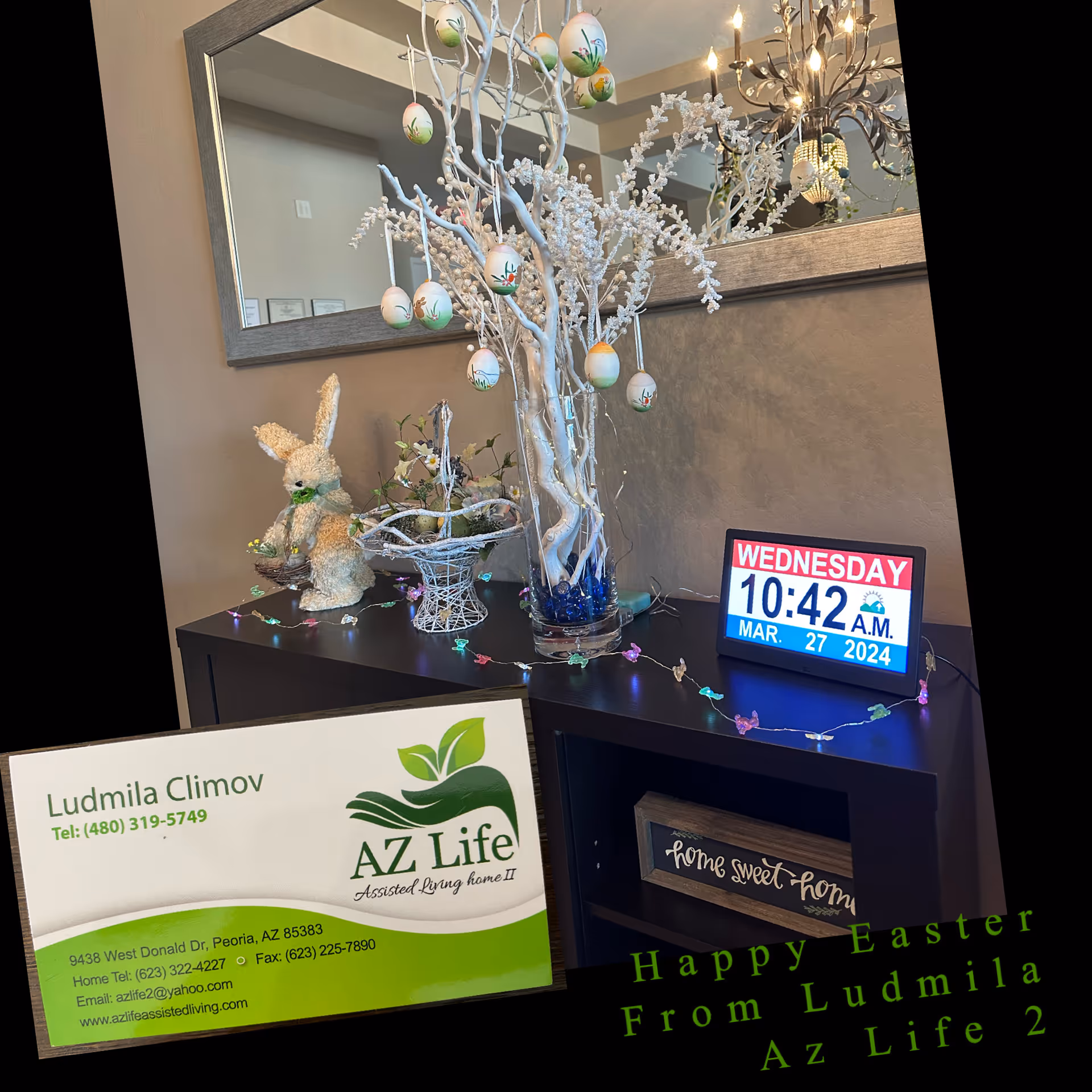 A decorated interior corner with a dark wooden cabinet topped with a white decorative tree adorned with hanging Easter eggs, a bunny figurine, a basket with flowers, and a digital clock displaying Wednesday, 10:42 A.M., March 27, 2024. A large mirror hangs on the wall above the cabinet, reflecting a chandelier. A small sign on the cabinet reads 'home sweet home'.