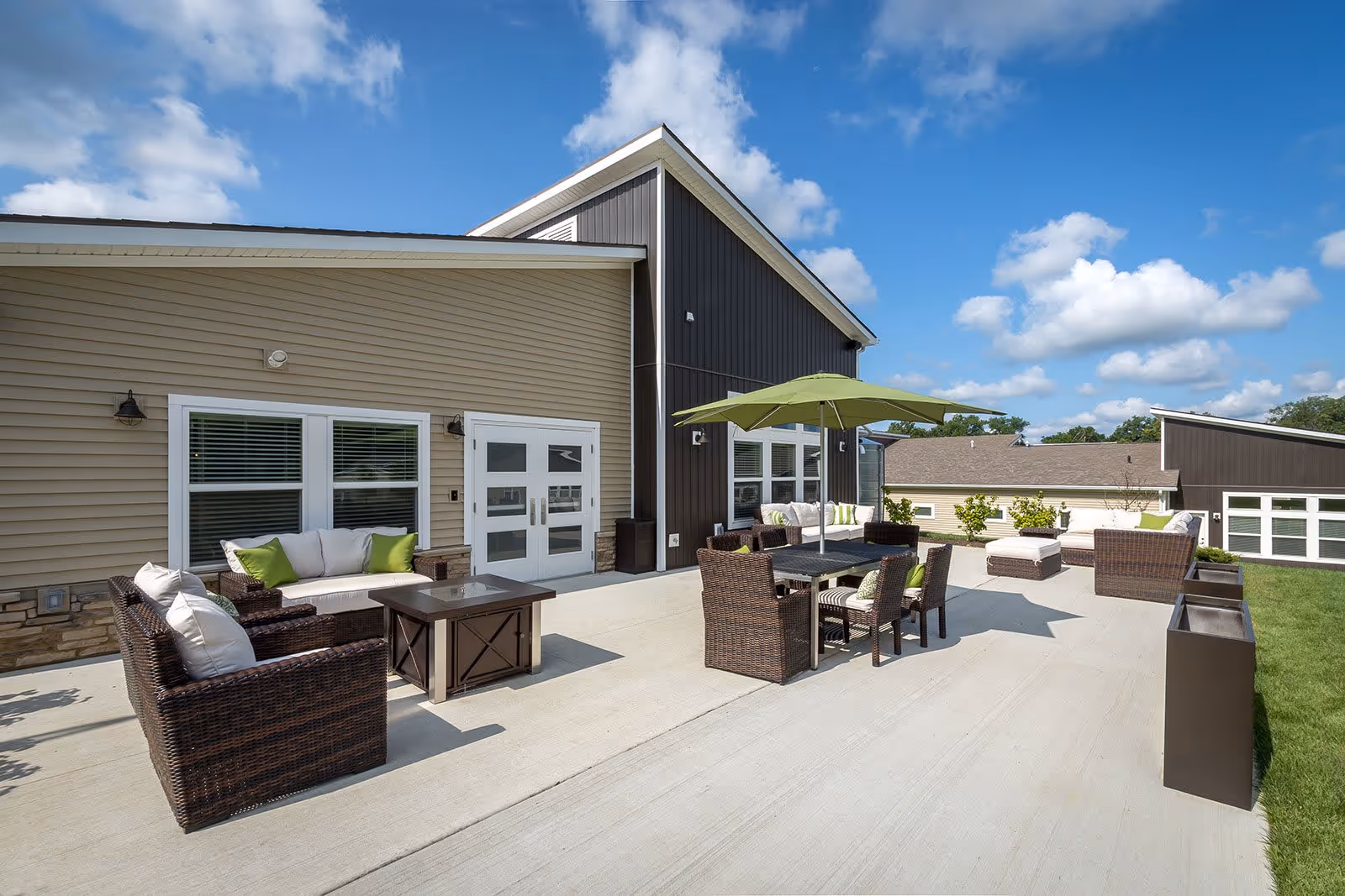 Outdoor patio area at Redbud Commons with wicker furniture including sofas, chairs, and tables. There is a green umbrella providing shade over one table. The patio is adjacent to a building with beige and dark brown siding under a blue sky with scattered clouds.