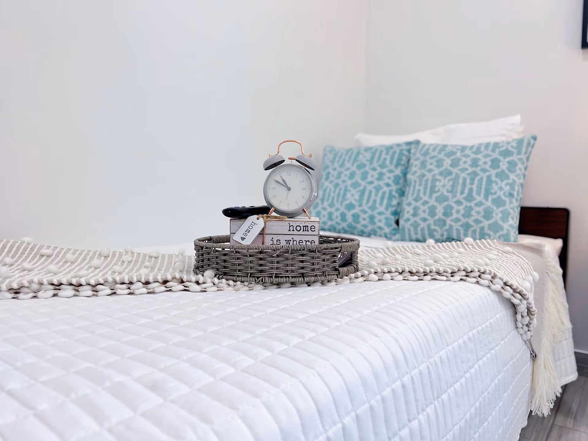 A neatly made bed with a white quilted bedspread and a beige textured throw blanket. On the bed is a round woven tray holding a small white alarm clock, a remote control, and decorative blocks with the words 'home is where'. Two blue patterned pillows and a white pillow are arranged against the headboard.