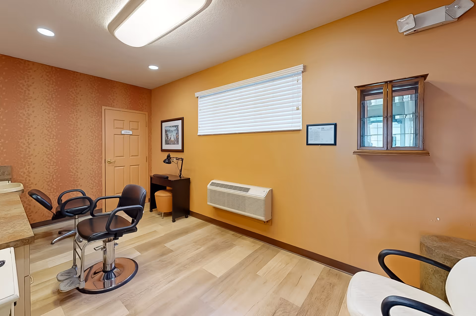 Interior room with orange walls and light wood flooring featuring two black salon chairs, a small desk with a lamp and stool, a wall-mounted air conditioning unit below a window with blinds, and a small wooden cabinet mounted on the wall.