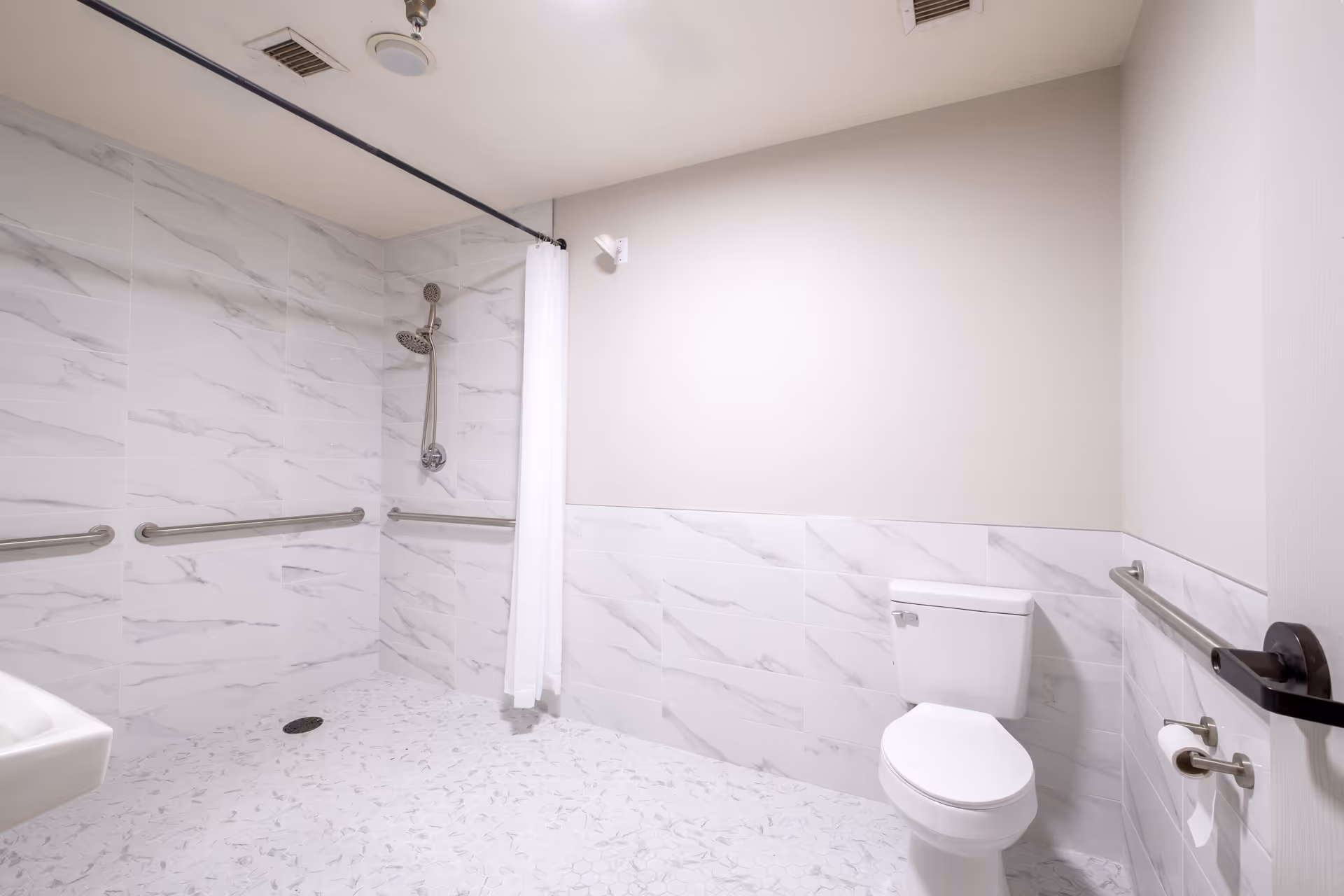 A clean, accessible bathroom with white marble-patterned tiles on the walls and floor. The bathroom features a walk-in shower with a handheld showerhead and a white shower curtain. There are multiple stainless steel grab bars on the walls for support. A white toilet is positioned near the door, which has a black handle. A small white sink is partially visible on the left side.