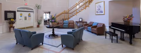 A spacious lobby with blue seating, a piano, and a staircase inside a senior living facility.