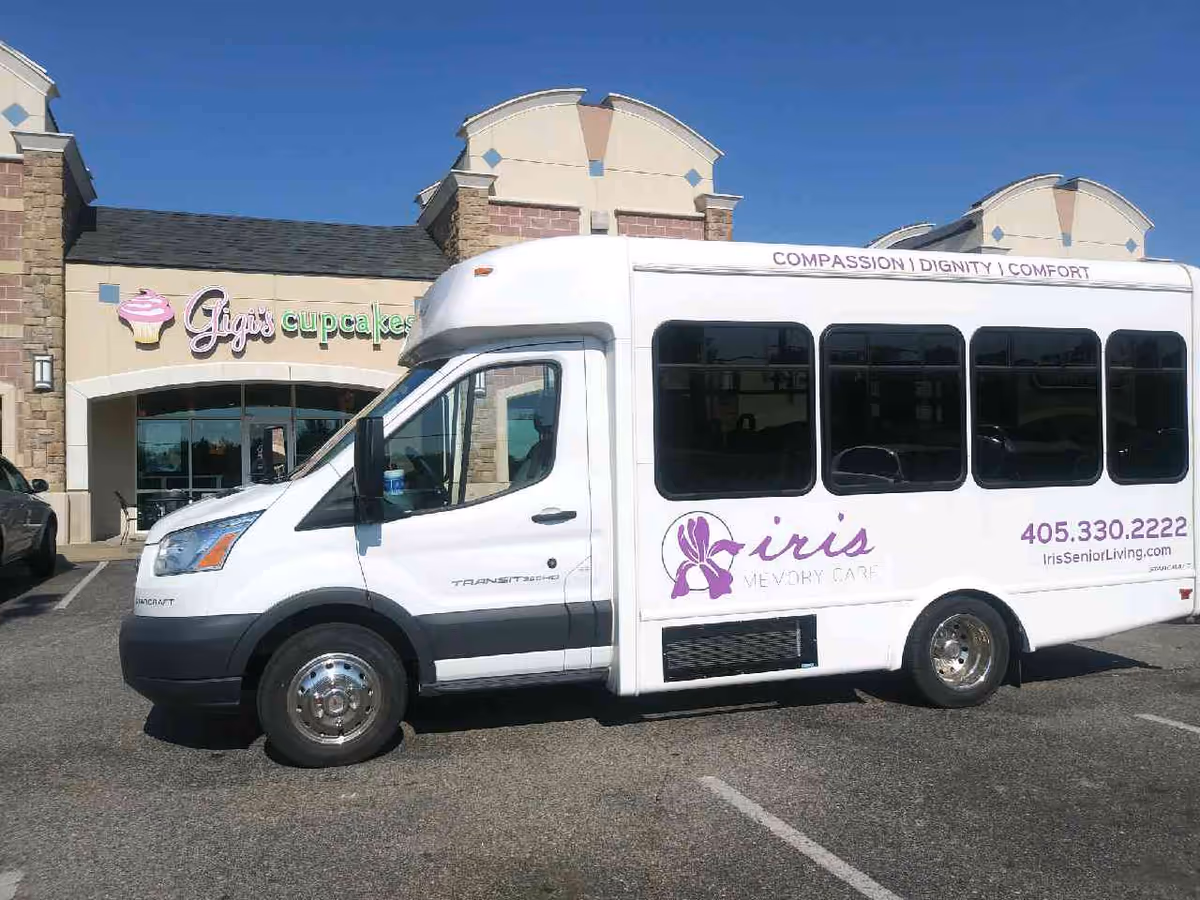 A white memory-care shuttle van parked in front of a shopping center storefront with a 'Gigi's cupcakes' sign.