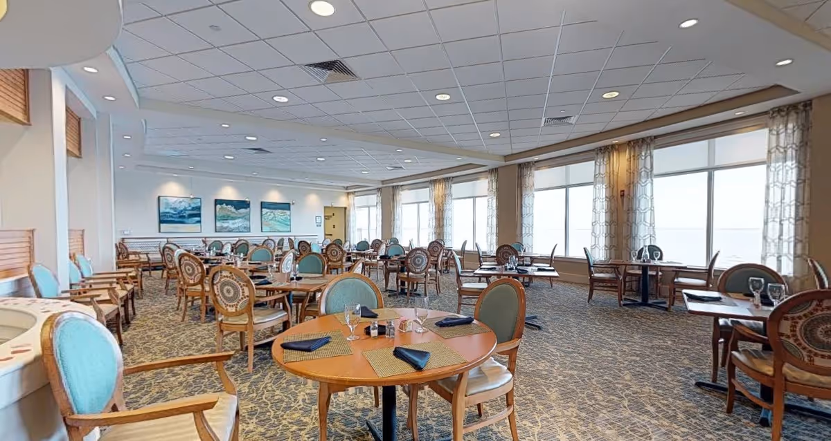 A spacious dining room with multiple round and square wooden tables set with placemats, napkins, and glassware. The room features large windows with patterned curtains allowing natural light to fill the space. The carpet has a patterned design, and there are several chairs with cushioned seats and backs arranged around the tables. Three paintings of ocean scenes hang on the far wall.