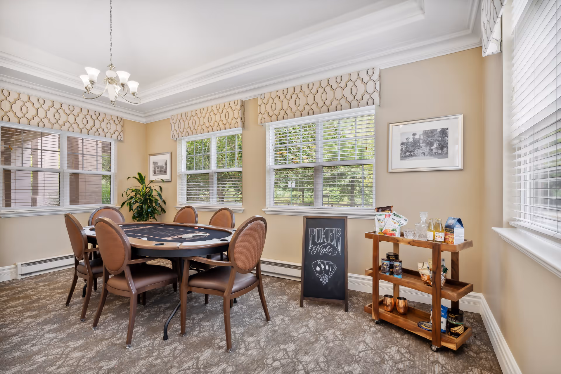 A well-lit room with a round poker table surrounded by six chairs. The room has three large windows with patterned valances, beige walls, and a chandelier hanging from a tray ceiling. There is a small wooden cart with snacks and drinks on the right side and a chalkboard sign that reads 'Poker Nights' with a playing card design. A potted plant and framed pictures are on the walls.