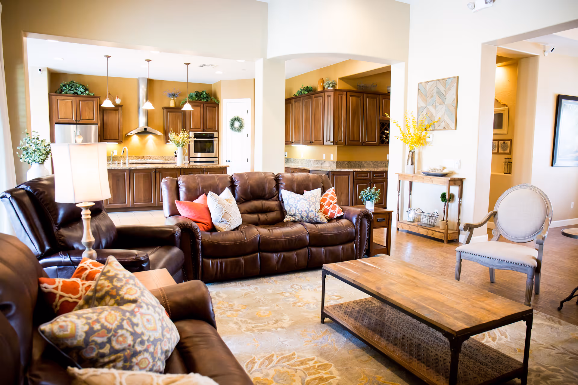 A bright and spacious living room with brown leather sofas adorned with colorful patterned pillows, a wooden coffee table, a beige upholstered armchair, and a lamp on a side table. In the background, there is an open kitchen with wooden cabinets, granite countertops, stainless steel appliances, and pendant lights hanging from the ceiling. The room has light-colored walls and wooden flooring, with decorative plants and artwork adding a cozy touch.