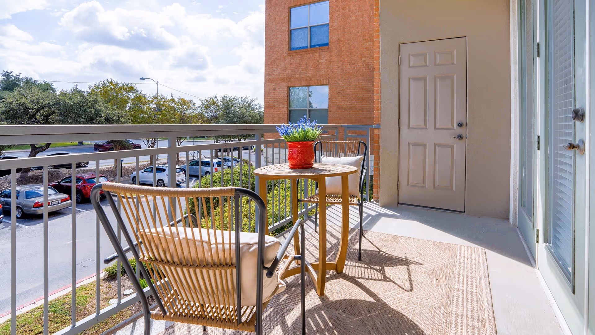 Balcony patio with a small round table, two chairs and a red potted plant overlooking a parking lot and trees.