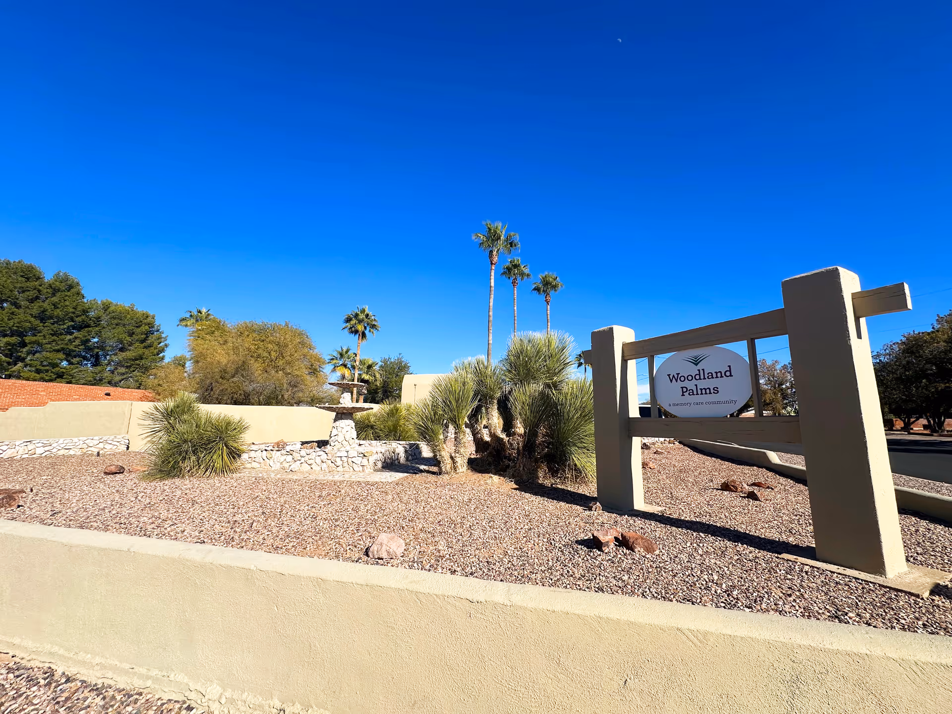 Entrance landscaping and sign for Woodland Palms memory care with palm trees under a clear blue sky.
