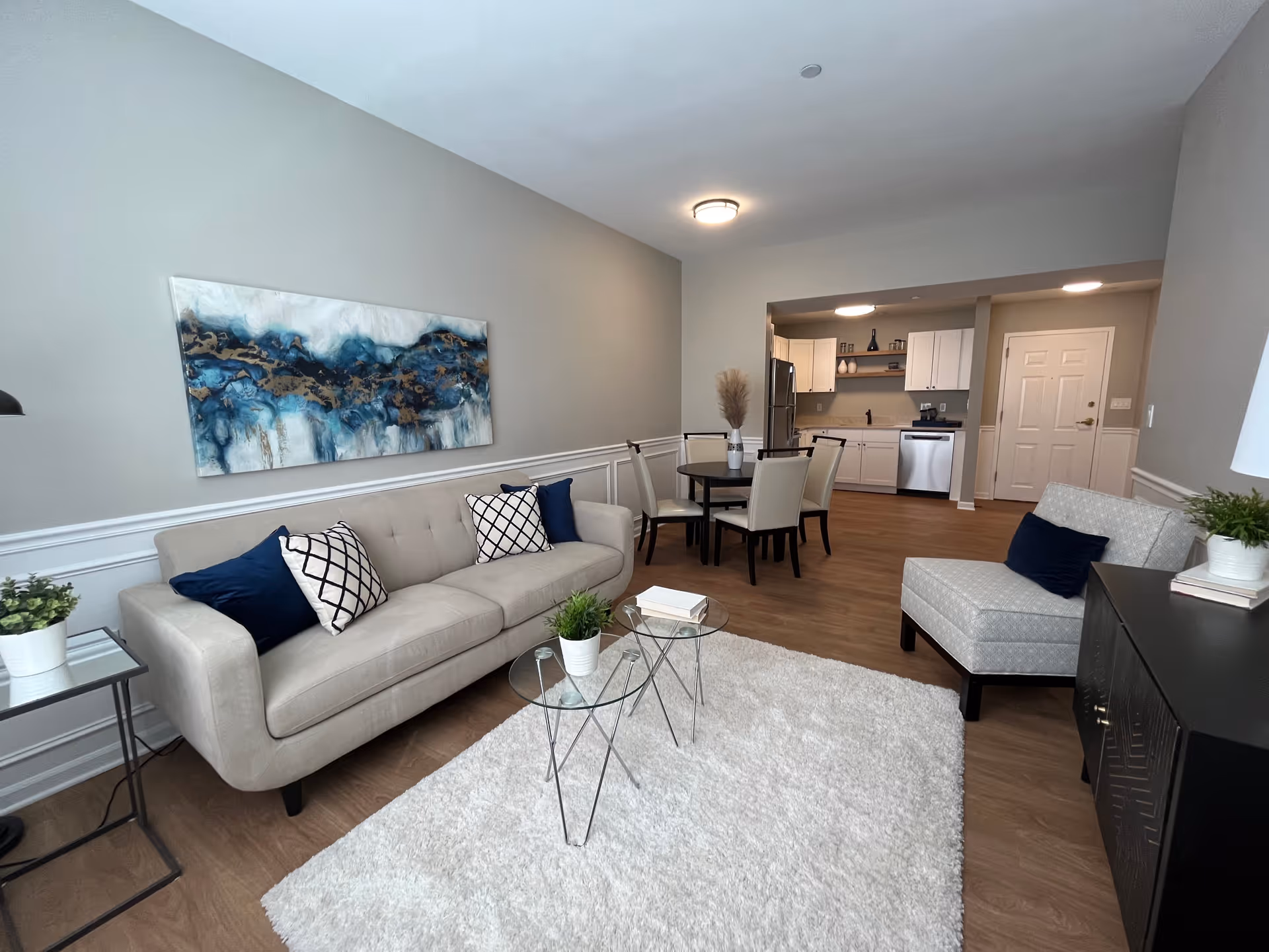 A modern living room and dining area with light gray walls and wood flooring. The living room features a beige sofa with blue and white patterned pillows, a matching armchair, two small glass coffee tables with plants and books, and a large abstract blue and gold painting on the wall. The dining area has a round table with four cream-colored chairs. In the background, there is a kitchen with white cabinets, stainless steel appliances, and a white door.