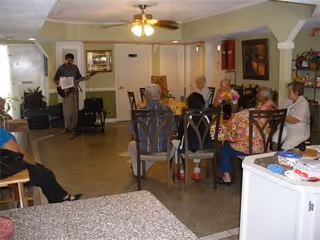 A group of elderly people seated around a table in a living room area, listening to a man playing an electric guitar. The room has a ceiling fan with lights, a painting on the wall, and various furniture including chairs and a countertop in the foreground.