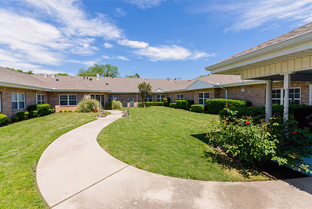 Curved concrete pathway leading through a well-maintained grassy courtyard surrounded by single-story brick buildings under a partly cloudy blue sky.