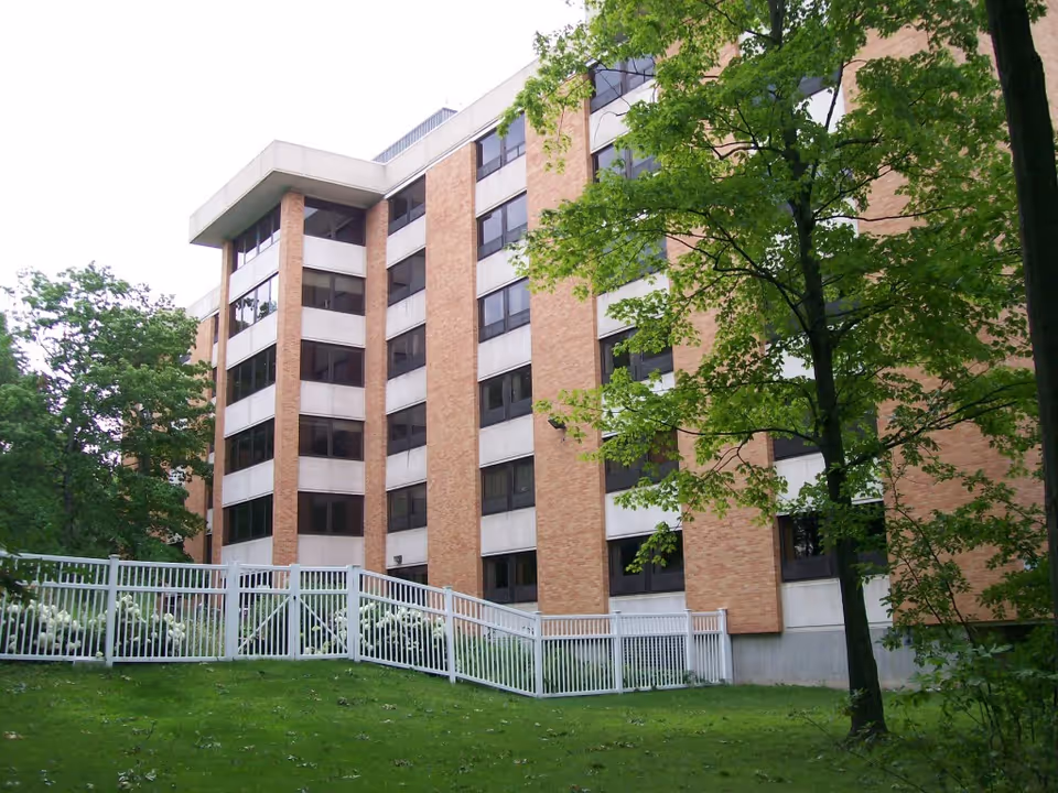 Exterior view of a multi-story senior apartment building with brick and concrete facade, surrounded by green trees and a white fence in the foreground.