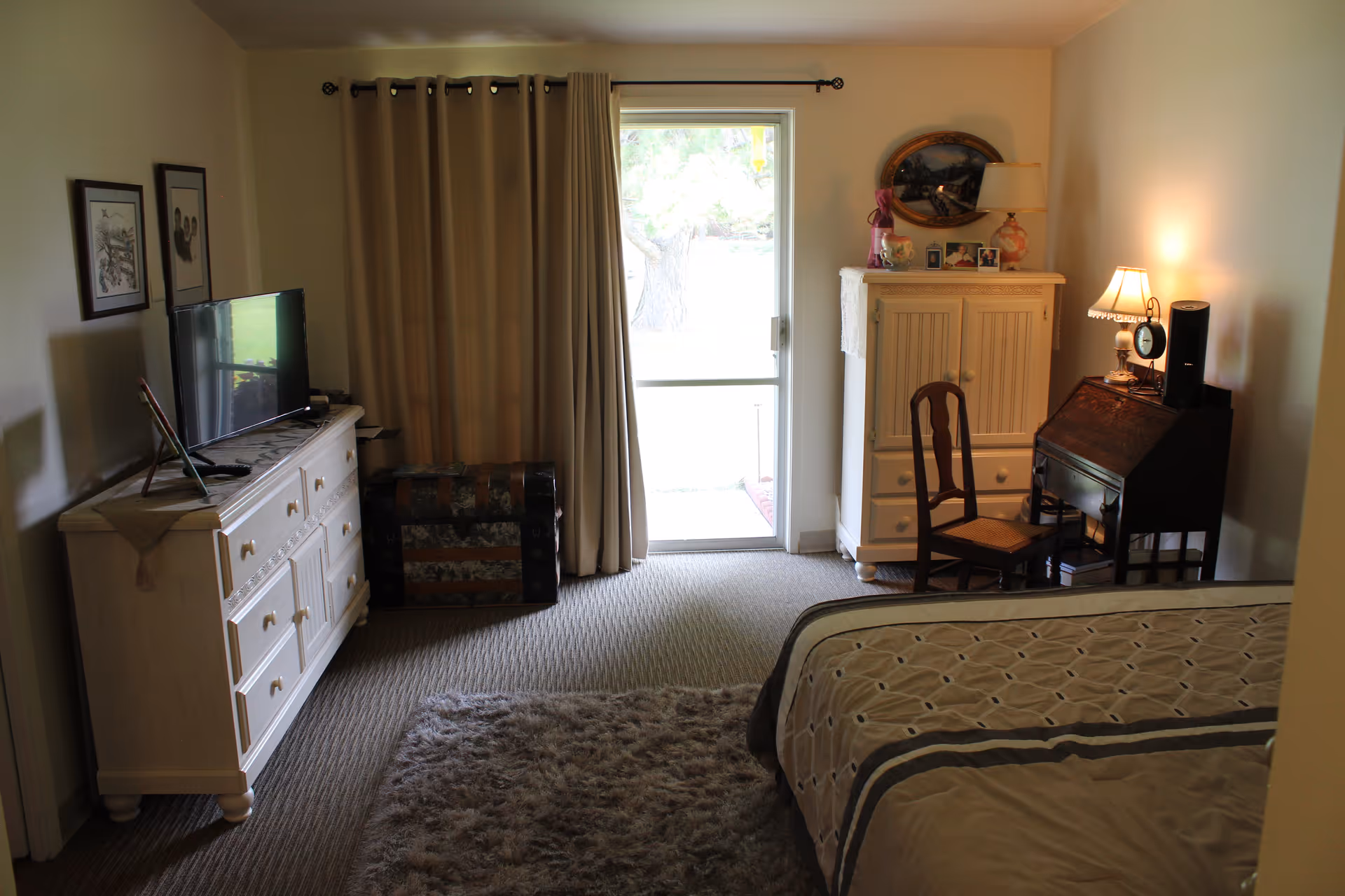 A cozy bedroom with a bed partially visible on the right, a dresser with a TV on the left, a trunk near a window with beige curtains, and a small wooden desk with a chair and lamp on the right side. The room has carpeted flooring and soft lighting.